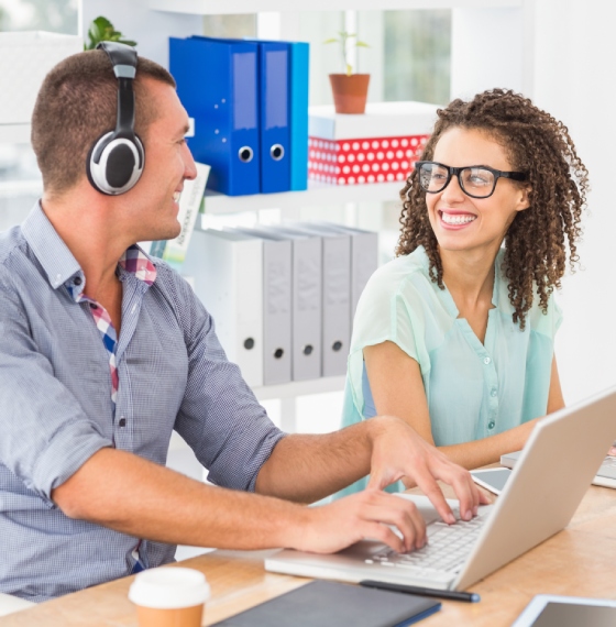 Muškarac sa slušalicama na ušima i žena sjede za radnim stolom i gledaju jedno u drugo dok kucaju na laptopu
